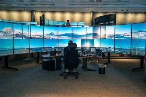image - The Travel Bloggers Control room with a curved wall of monitors displaying a panoramic landscape; a technician sits at a desk with multiple computer screens.